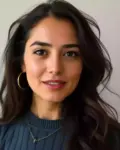 Portrait of a young woman with long dark hair, gold hoop earrings, and a blue knit top smiling softly.